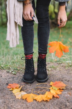 Gri ceketli, bej atkılı ve siyah kotlu şık beyaz kadın kahverengi deri çanta ve turuncu akçaağaç yaprağı tutuyor. Arka planda sonbahar manzarası. Kamu parkında yürüyorum. Düşen yapraklar