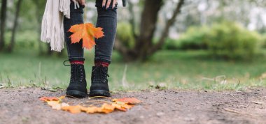 Gri kotlu, kırmızı örgü çoraplı, bej eşarplı ve elinde turuncu akçaağaç yaprağı tutan siyah çizmeli modaya uygun beyaz kadın. Sarı yapraklar kızdan önce gelir. Arka planda yeşil çimen. Metin için boşluk.