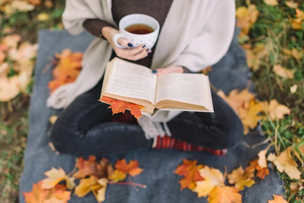 Siyah kotlu, kahverengi kazaklı, bej renkli kitap okuyan ve çay içen bir kadın. Ilık içeceklerle dolu bir fincan. Düşmüş akçaağaç yaprakları ayak altında. Kitabın içindeki yaprağa odaklan. Sonbahar havası.