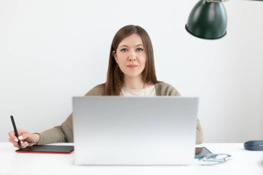 Çekici grafik tasarımcısı ya da rötuş makinesi masada dizüstü bilgisayarla oturuyor ve kameraya bakıyor. Gri hırkalı kız grafik tablet için el kalemi tutuyor. Seçici bir şekilde kadınlara odaklan. Kapat..