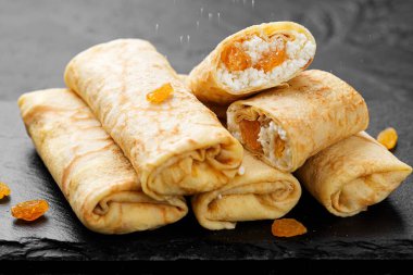 Süzme peynirle doldurulmuş ince krepler ya da krepler ve siyah arka planda kuru üzümler. 