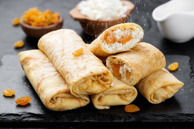 Süzme peynirle doldurulmuş ince krepler ya da krepler ve siyah arka planda kuru üzümler. 