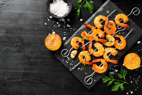 Grilled shrimp skewers. Shrimps with herbs, garlic and lemon on black background.