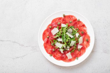 Carpaccio bifteği, parmesanlı soğuk aperatif, kapari ve roka beyaz tabakta..
