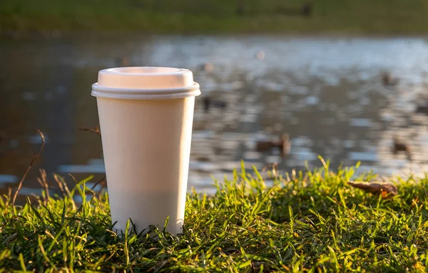 A white glass for a drink stands on the green grass against the backdrop of the lake.