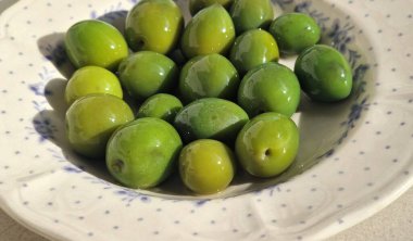 Taze yeşil zeytinler güneş ışığında parıldıyor, klasik desenli bir tabakta sıralanmış.