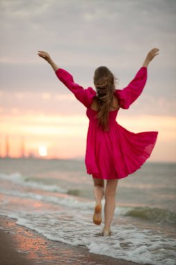 Young girl in a pink summer dress walks on the sea coast at sunrise. Blurred photo. Out of focus. clipart