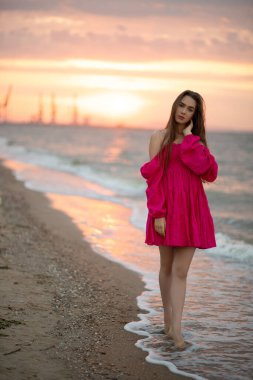 Pembe elbiseli genç kız gün doğumunda deniz kıyısında yürüyor.