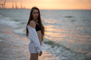 Beyaz gömlekli genç kız gün doğumunda deniz kıyısında yürüyor.