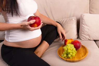 Hamile bir kadın elinde bir tabak meyveyle kanepede oturur ve elinde şeftali tutar. Yüksek kalite fotoğraf