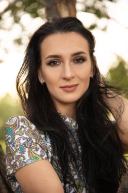 Koyu renk saçlı güzel bir kadının portresi, yaz günbatımının arka planında bir kız ve yeşil bir park.