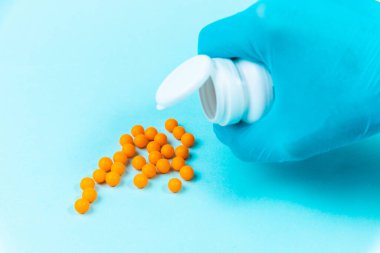 Mavi eldivenli bir el mavi arka plandaki kavanozdan sarı haplar döker..