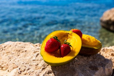 Olgun mango ve çilekler denize karşı bir taşın üzerinde yatıyor.
