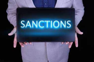 SANCTIONS metin, kelime siyah bir laptop üzerinde neon harflerle yazılmış, Gri takım elbiseli bir işadamının önünde.