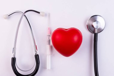 Bir stetoskop, bir şırınga ve kırmızı bir kalp beyaz arka planda yatıyor. Sağlık, kardiyoloji ve medya kavramı.