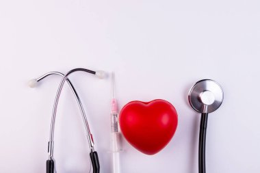 Bir stetoskop, bir şırınga ve kırmızı bir kalp beyaz arka planda yatıyor. Sağlık, kardiyoloji ve medya kavramı.