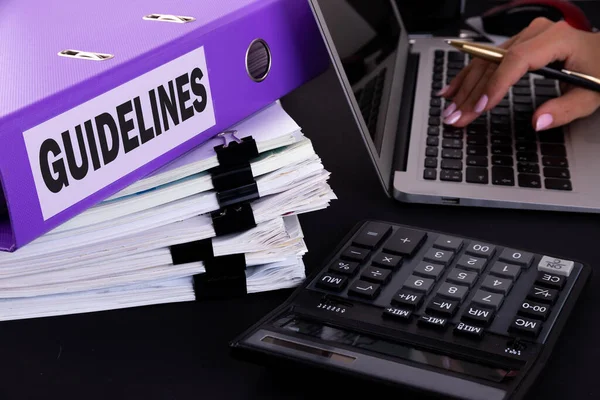 Üzerinde Kelime Kılavuzu yazan dizin, karanlık bir ofis masasında dizüstü bilgisayarlı, kalemli, hesap makineli ve raporlu.