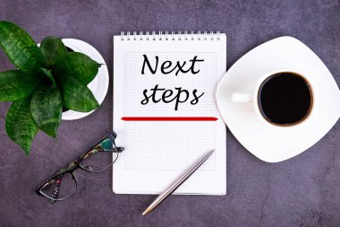 Not defteri olan ofis masası. Kalemle, bardaklarla ve bir fincan kahveyle NEXT STEPS yazıldı..