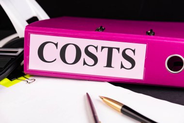 Metin KOSTS bir klasör üzerine yazılır. Masada bir kalem ile bir yığın kağıt üzerinde yatar. İş konsepti.