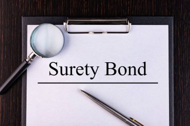 Text SURETY BOND is written on a notebook with a pen and a magnifying glass lying on the table. Business concept. clipart