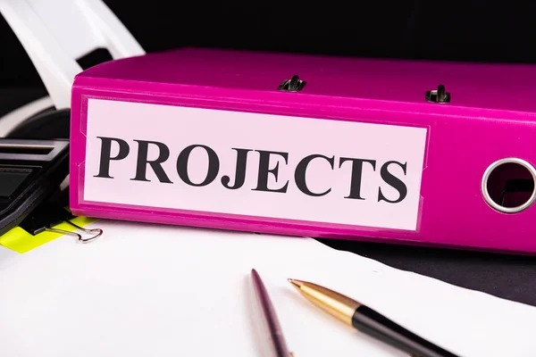 Text PROJECTS is written on a folder lying on a stack of papers with a pen on the table. Business concept.