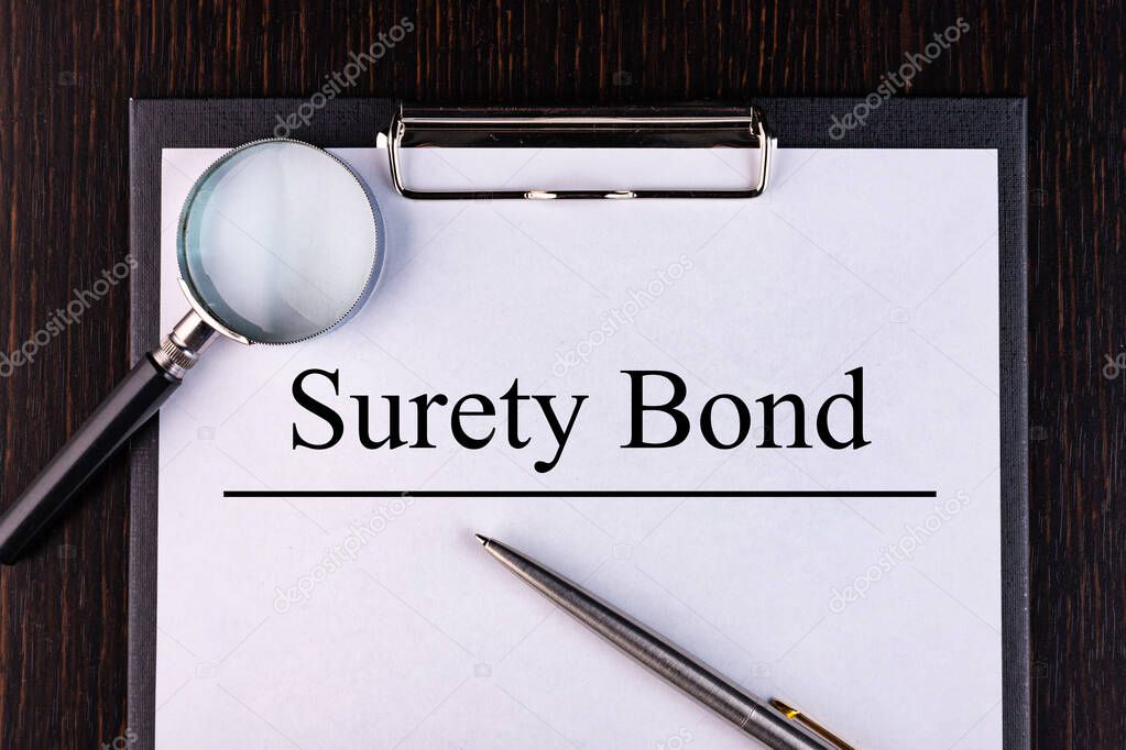 Text SURETY BOND is written on a notebook with a pen and a magnifying glass lying on the table. Business concept.