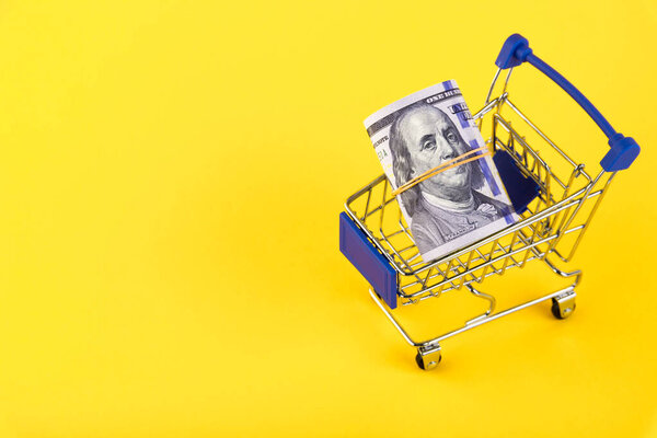 Корзина, тележка супермаркета с рулоном стодолларовых купюр, на желтом фоне. Концепция получения кредита. Копирование пространства