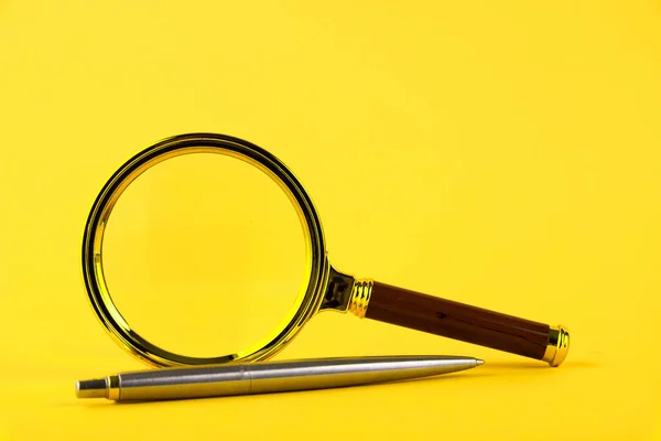 A magnifying glass in a gold frame and a pen on a yellow background. Copy space.