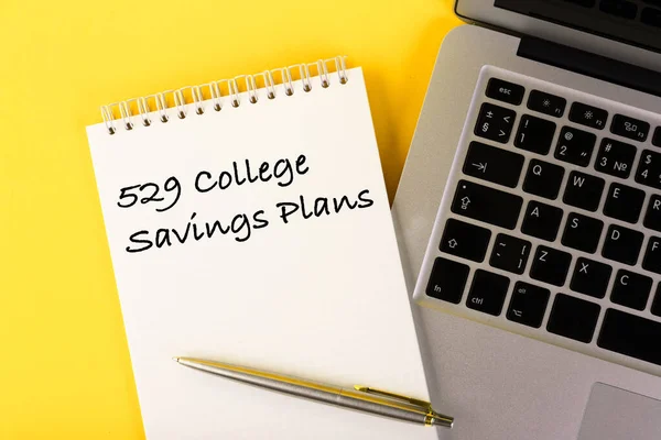 Text, 529 College Savings Plans, Education Loan, written in a notebook lying on a desk with a notebook on a yellow background.