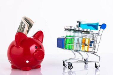 Bir alışveriş arabasındaki aşı için farklı renklerde cam şişeler, beyaz arka planda para için kırmızı domuz kumbarası. Uzayı kopyala Seçici odak.