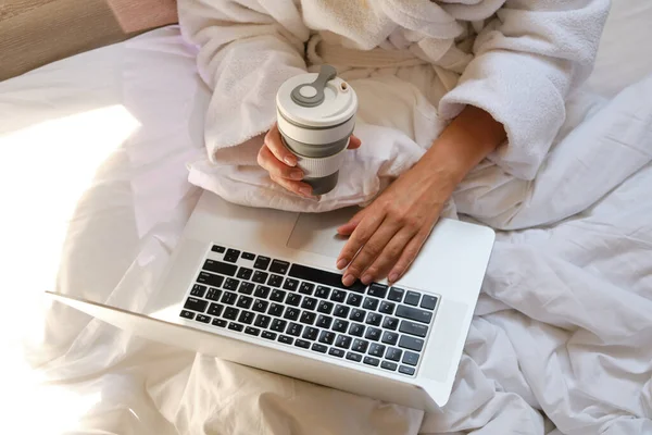 Woman with laptop and coffee cup in bed. Girl in white bathrobe. Drinking morning coffee and work online in home. Cozy relaxation in bed. Distance freelance work. Technology in lifestyle, new reality