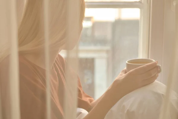 这个女孩早上在窗边喝茶 手里拿着一杯热茶 从睡眠中醒来 这个快乐的女人裹在毛毯里 想想生活 在家里享受舒适吧 — 图库照片