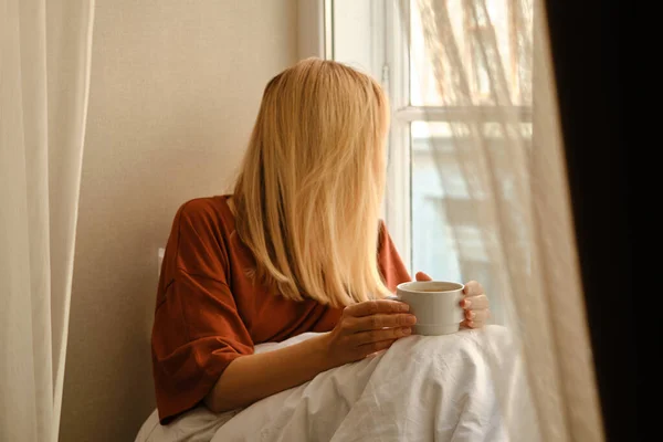 The girl drinks tea in the morning near the window. A cup of hot tea in hands. Awakening from sleep. The happy woman is wrapped in a blanket. Dreams and think about life. Enjoy cozy in home