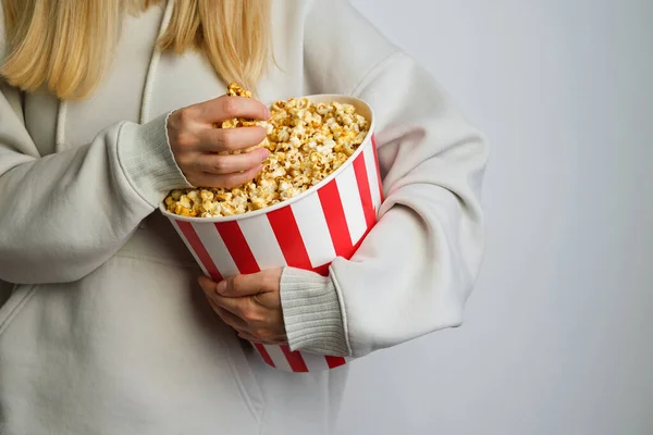 Popcorn paper bucket in the hands of a young girl preparing to watch a movie. Showtime. Eating delicious unhealthy sweet snacks. Going to cinema for a new film. Rest and entertainment. Popcorn closeup