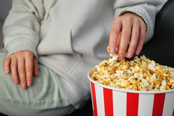 Popcorn paper bucket in the hands of a young girl preparing to watch a movie. Showtime. Eating delicious unhealthy sweet snacks. Going to cinema for a new film. Rest and entertainment. Popcorn closeup