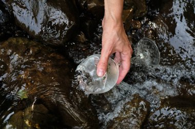 Gezgin kız bir dağ nehrinden soğuk suyu bardağa boşaltıyor. Tazelik sıçrıyor. Şarap kadehindeki doğal su. Sıcak yaz gününde dinlenme. Sağlıklı yaşam tarzı, sağlık. Doğayla bütünleşen kadın