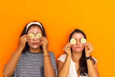 Turuncu arka planda gözlerine salatalık parçaları tutarken güzellik maskeli iki güzel Latin kadın. Ev kaplıcası