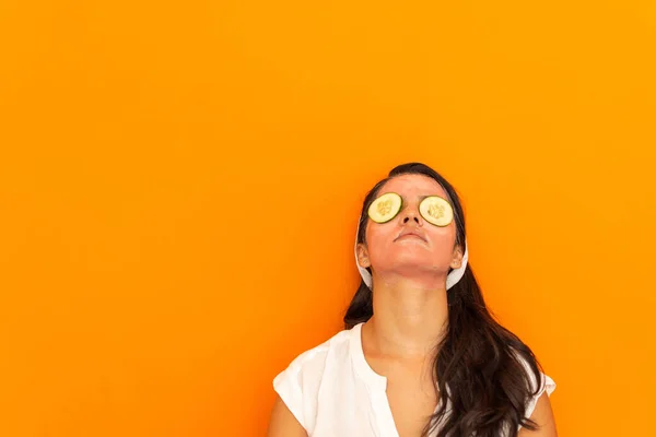 Yüzünde güzellik maskesi, gözlerinde turuncu bir arka planda salatalık parçaları olan genç ve güzel bir Latin kadın. Kişisel bakım kavramı