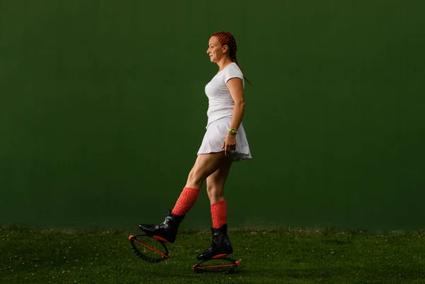 Beyaz spor giyim ve kangoo çizme yan duran Latin kadın, koyu yeşil bir arka planda, bir bacağını öne koyuyor