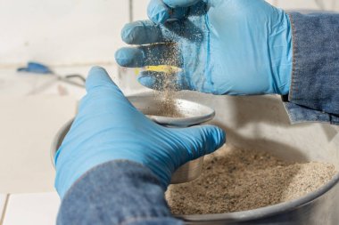Mavi lateks eldivenli erkek sanayi işçisinin elleri metal bir kaba kum döküyor.