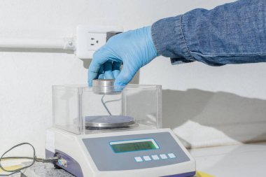 Mavi lateks eldivenli işçi el, metalik bir parçayı tartıya koyuyor.