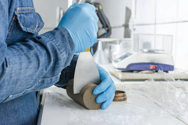 Mavi lateks eldivenli endüstriyel işçi elleri bir spatulayla kil silindiri kesiyor.