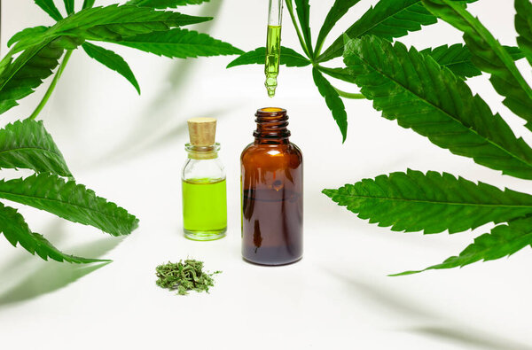 Медицинские бутылки с марихуаной, окруженные листьями марихуаны на белом фоне, в то время как заполненная маслом капельница бросает каплю в одну из бутылок