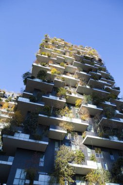 Milano 'daki Bosco Verticale (Dikey Orman) yerleşim kulelerinin şaşırtıcı düşük açılı görüntüsü, açık mavi gökyüzü altında çeşitli ağaçlar ve bitkilerle entegre yemyeşil balkonların yer aldığı.