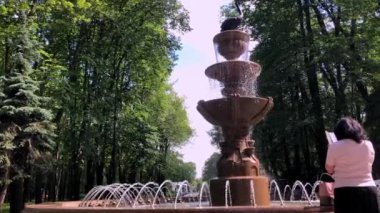 Sırtı dönük bir kadın şehir parkında bir cep telefonu fıskiyesine takılıyor. Yaz çeşmesinden su akar..