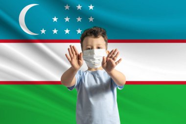 Özbekistan bayrağının arka planında koruyucu maskeli küçük beyaz çocuk. Elleriyle dur işareti yapıyor, Özbekistan 'da kalıyor..