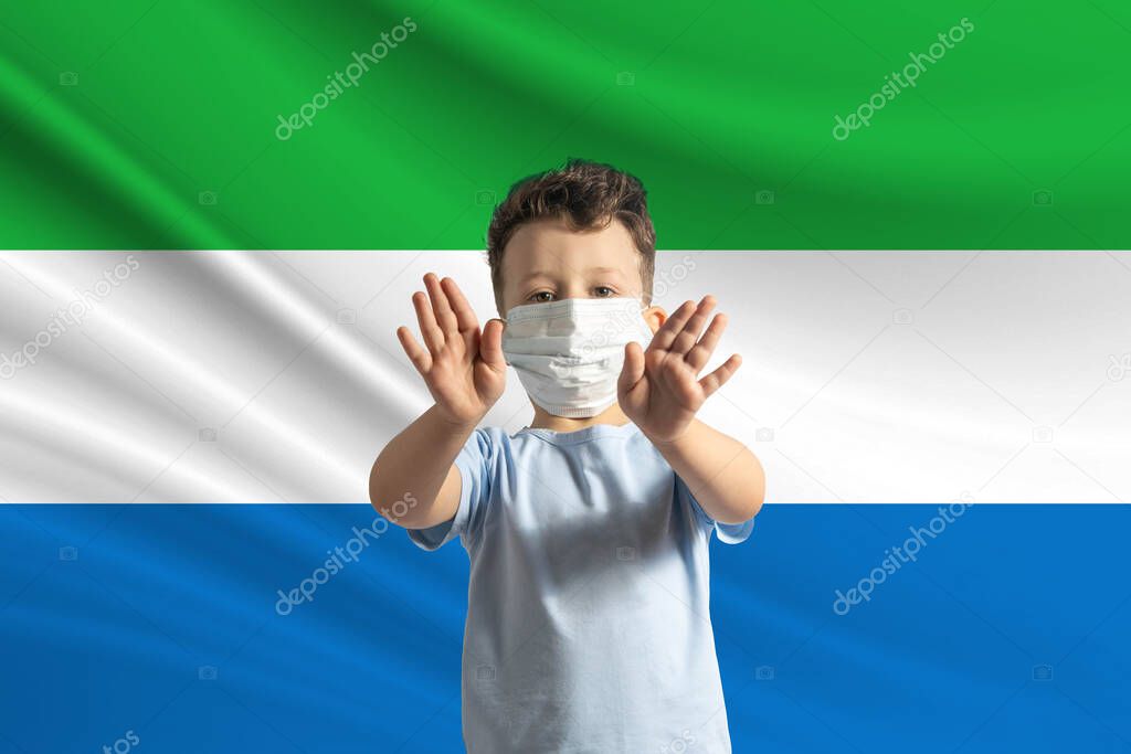 Little white boy in a protective mask on the background of the flag of Sierra Leone. Makes a stop sign with his hands, stay at home Sierra Leone.