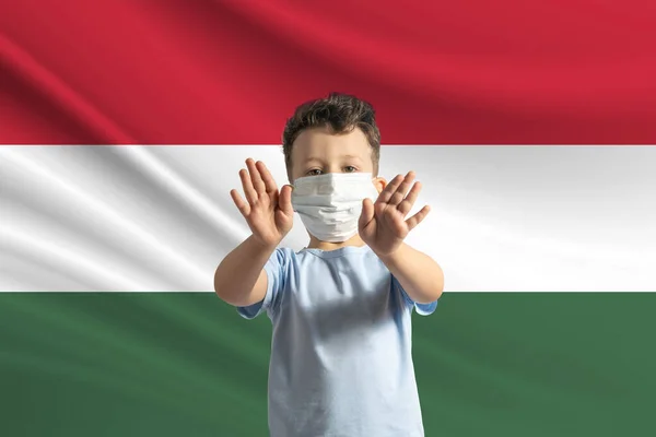 戴着保护面具的白人小男孩站在匈牙利国旗的后面 用手做一个停车标志 呆在家里 — 图库照片