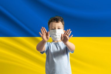 Ukrayna bayrağının arka planında koruyucu maskeli küçük beyaz çocuk. Elleriyle dur işareti yapar, Ukrayna 'da kalır..