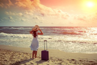 Hasır şapkalı, seyahat valizli genç ve mutlu bir kadın yazın sahilde yürüyor. Kadın hasır şapkayı tutuyor ve denize bakıyor. Seyahat, iyi tatiller, yaz tatili konsepti